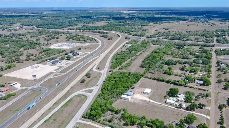 Wide aerial view of a highway interchange with frontage roads, wooded acreage and nearby buildings under a clear sky