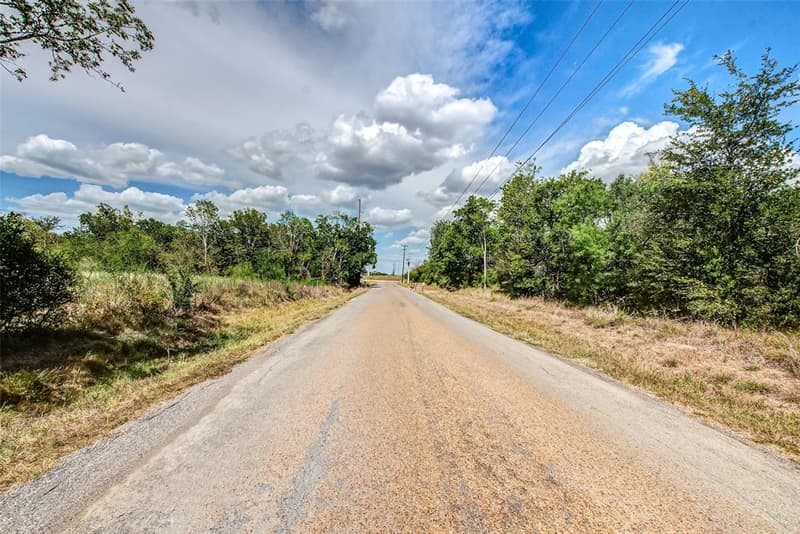 Paved two-lane road lined with trees and tall grass under a blue sky with white clouds