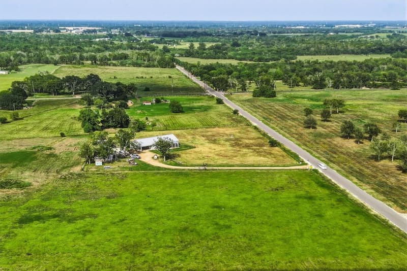 Aerial view of the fenced acreage with a gray metal-roof barndominium, a long driveway and a paved country road