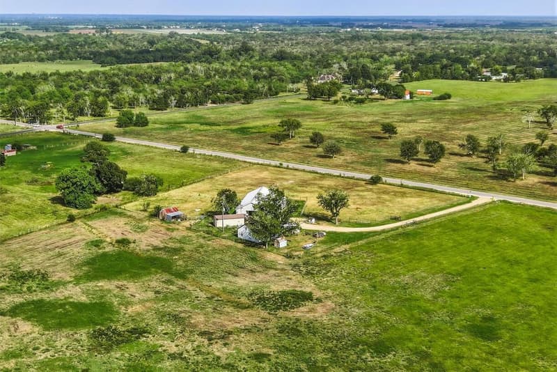 Aerial view of the barndominium, a white second home and outbuildings, with open pasture and trees around them