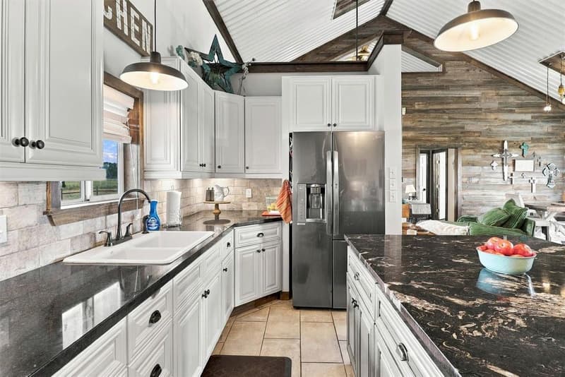 Kitchen with white cabinets, dark granite counters, a white sink under a window and a stainless refrigerator