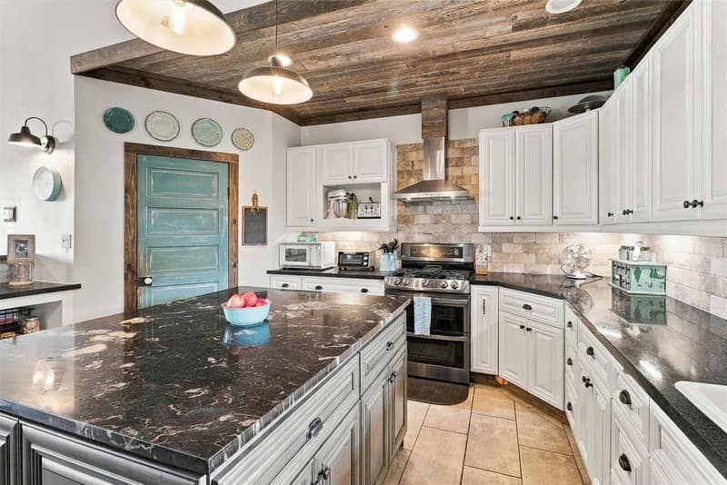 Kitchen island with dark veined granite, a stainless gas range with vent hood, stone-look backsplash and teal door
