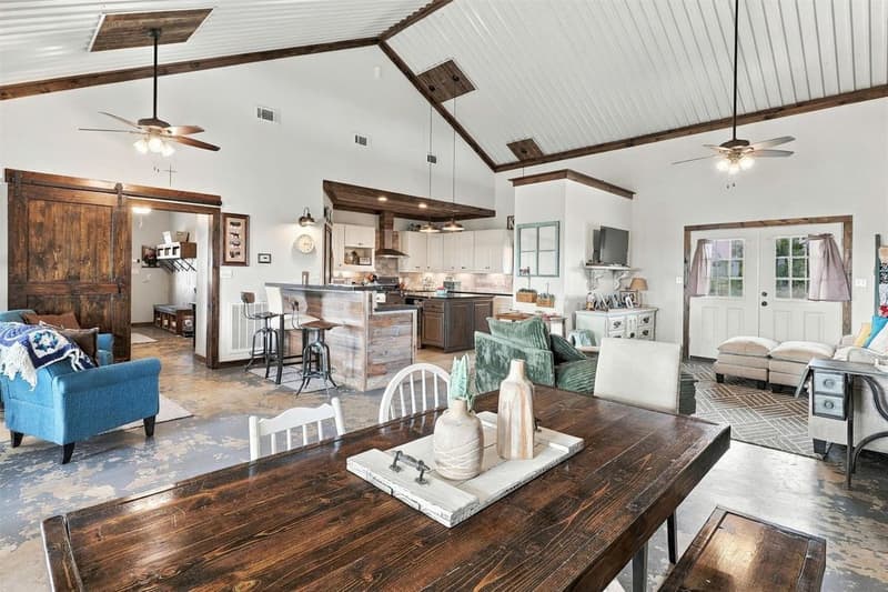Open great room with vaulted metal ceiling and wood beams, dining table, sliding barn door and kitchen with bar-height island
