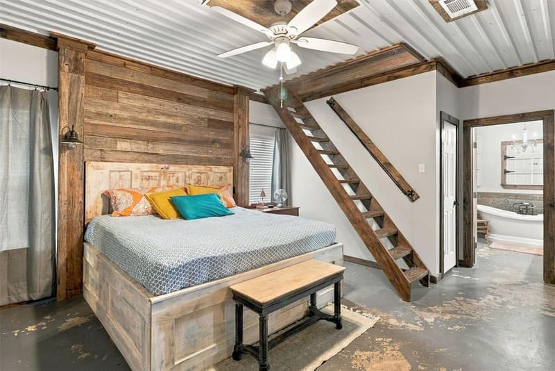 Primary bedroom with wood plank accent wall, ladder stair with iron handrail, ceiling fan and view into the en-suite bath