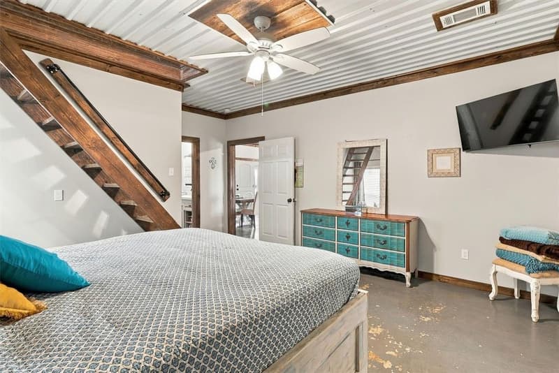 Primary bedroom seen toward the doorway, with wall-mounted TV, painted dresser, ceiling fan and ladder stair at left