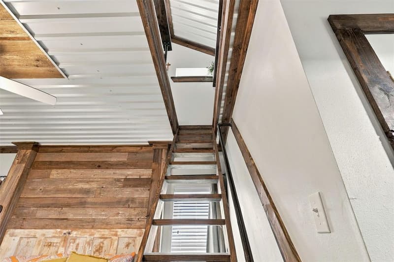 Looking up a wooden ladder stair from the bedroom to the loft, framed by stained wood beams and a metal ceiling