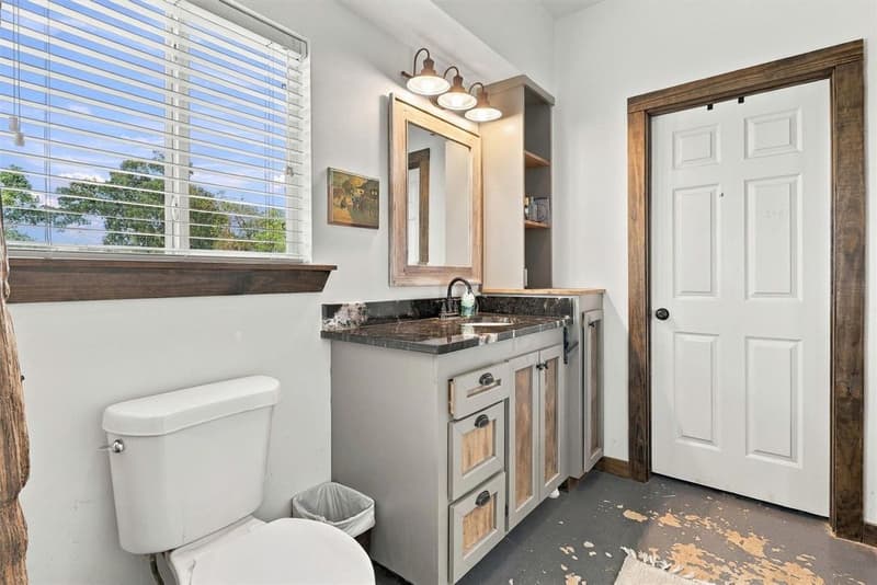 Bathroom vanity with dark stone counter, barn-style light bar, open shelving and window with a view of trees