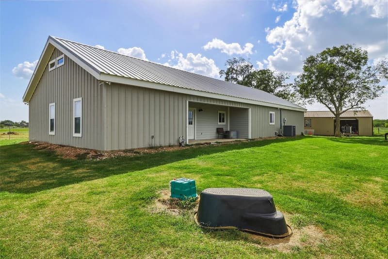 Gray metal-sided barndominium with a metal roof and covered porch, green lawn and a detached shed under large trees