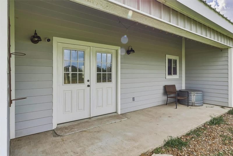 Covered concrete porch with white double glass-pane doors, barn-style wall lights and light gray siding