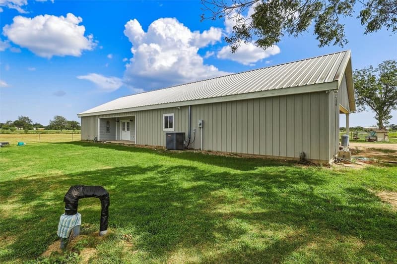 Long side of the gray metal barndominium with metal roof, covered entry porch, AC unit and a mowed lawn under blue sky