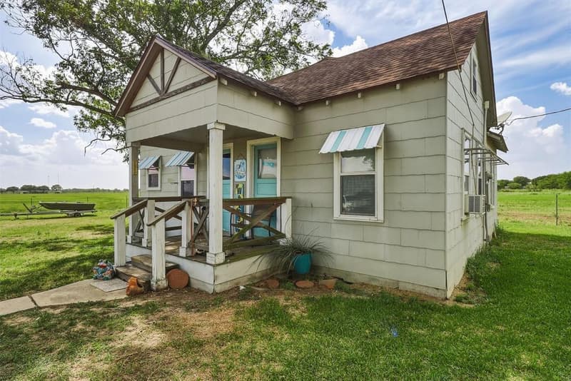 Small gray cottage with a covered wooden front porch, striped window awnings and a turquoise front door under a large tree