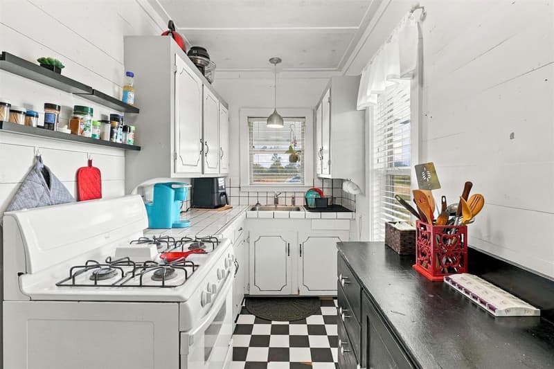 Galley-style cottage kitchen with white cabinets, white gas range, tile counters and black-and-white checkered floor