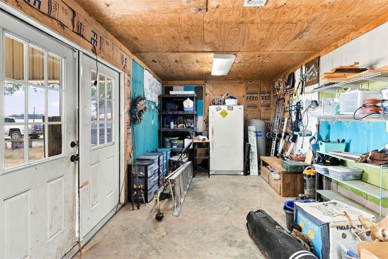 Enclosed workshop room with plywood ceiling, concrete floor, shelving, a freezer and stored tools and supplies