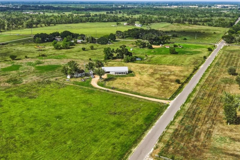 High aerial drone view of the property with its driveway off a paved country road, surrounded by open pasture