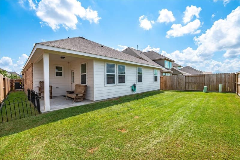Fenced backyard with wide lawn and covered back patio with wooden chairs along the rear of the home