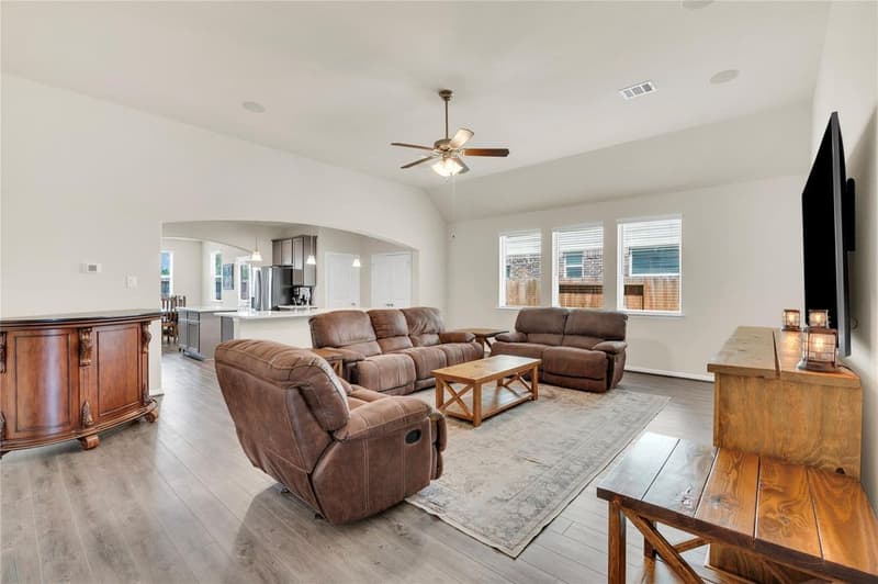Open family room with ceiling fan, three windows and recliner seating, open to the kitchen through a wide arch