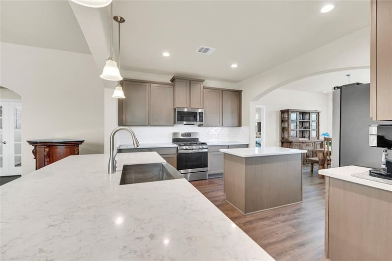 Quartz island with stainless sink facing a second island, gas range and shaker cabinets with subway tile backsplash
