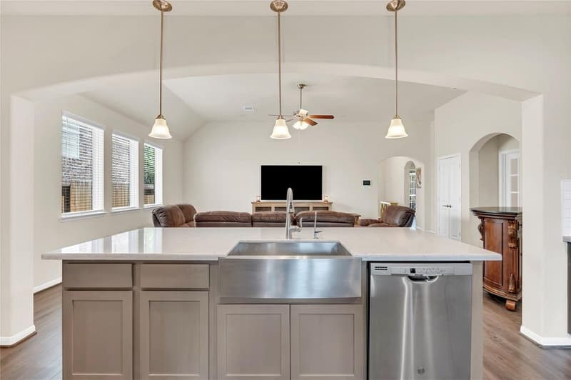 Island with a stainless farmhouse sink and dishwasher under three pendant lights, looking into the family room