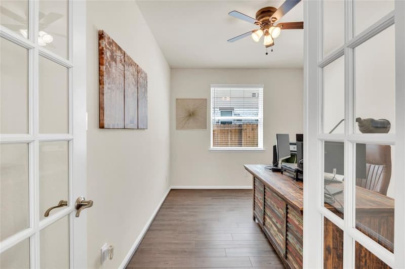 Home office seen through glass-paneled French doors, with window, ceiling fan and vinyl plank flooring