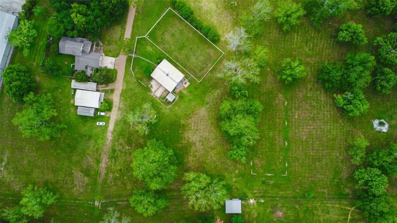Top-down drone view of a rural property with a house, outbuildings, a horse barn beside a white-fenced paddock, a long drive and scattered trees