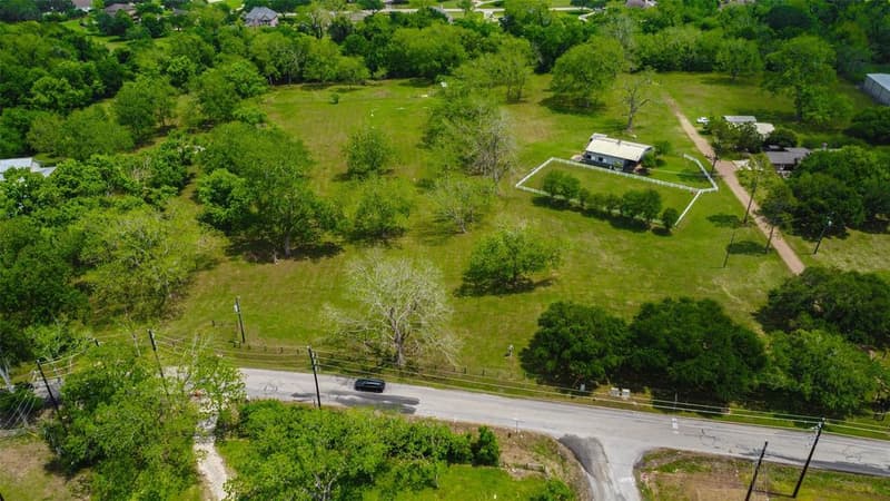 Drone view from the road of a tree-dotted pasture with a white-fenced paddock, a barn and a gravel drive leading back to buildings