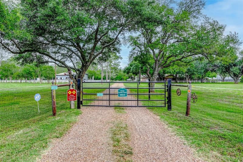 Black metal ranch gate on a gravel drive between wire-fenced pastures, under large shade trees, with address 2810 on the gatepost