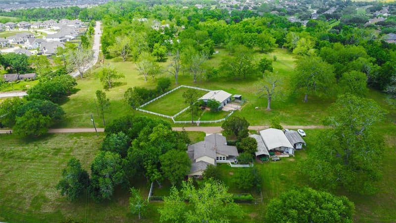 Drone view of a ranch-style home with outbuildings and carports, a horse barn with white-fenced paddock and wooded acreage behind