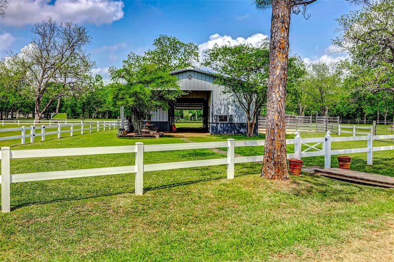 Gray metal horse barn with an open center aisle, set behind white two-rail fencing on green pasture under shade trees