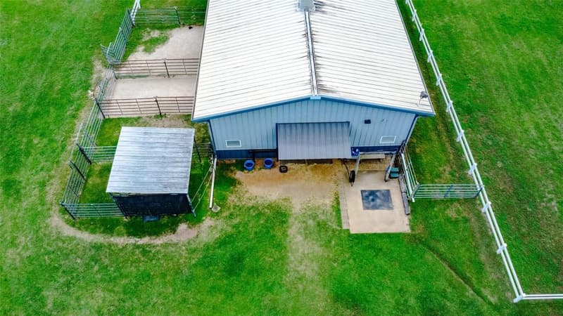 Overhead drone view of a metal barn roof with pipe-fenced pens, a small covered shed and a concrete pad beside white fencing