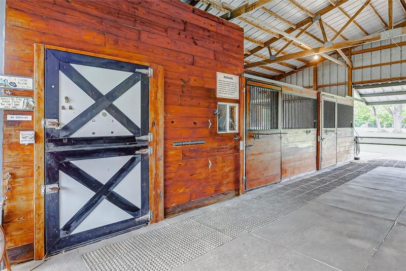 Barn aisle with wood-paneled walls, a dutch door with X-braced panels, stall fronts with metal grilles, and a rubber-mat walkway