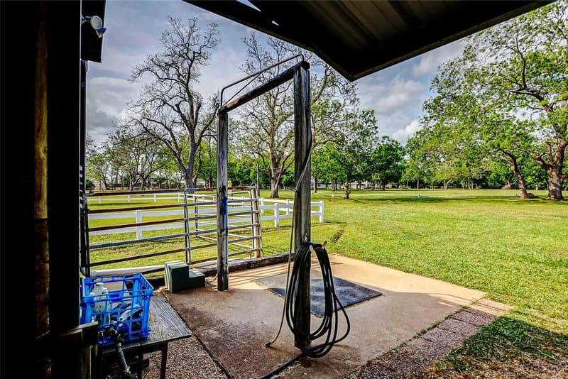 View from under a barn overhang of a concrete wash pad with hose and tie post, pipe gate, white fence, and a pasture with large trees