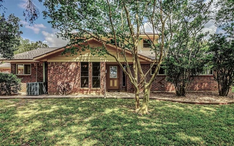 Two-story red brick ranch-style home with a gabled entry, glass-paned door, wagon wheel decor, and shade trees on a green lawn