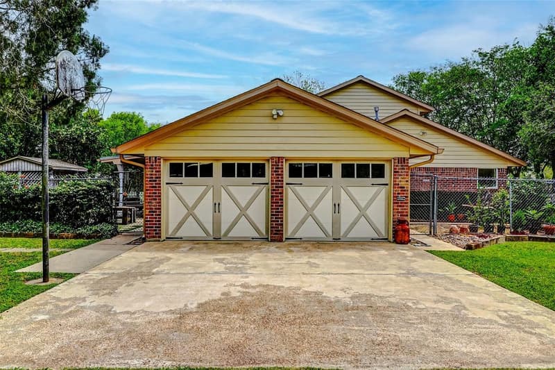 Garage with two cream carriage-style doors and brick columns, a wide concrete driveway, a basketball hoop, and chain-link fencing