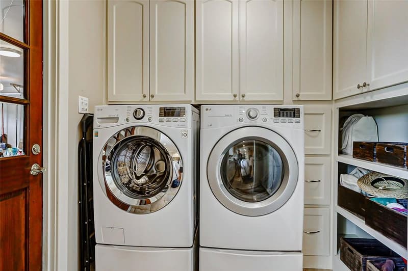 Laundry room with a front-load washer and dryer on pedestals, cream cabinets above, drawers, and open shelves with baskets