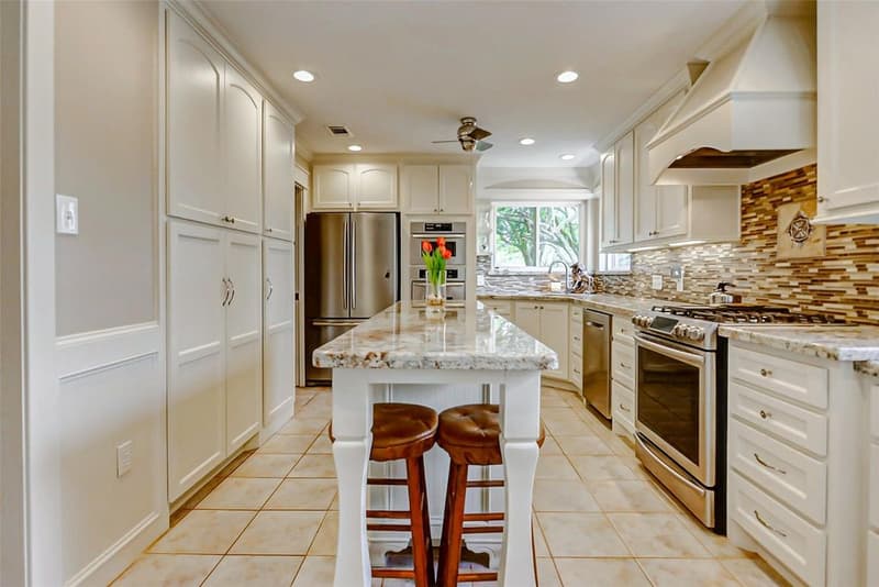 Remodeled kitchen with cream cabinets, a granite island with two stools, stainless appliances, a gas range, a vent hood, and a tile backsplash