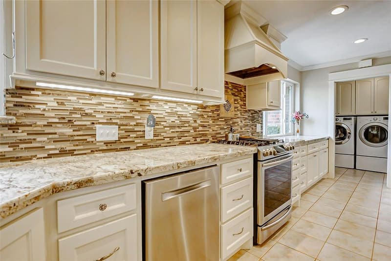 Kitchen counter with granite tops, a glass-and-stone mosaic backsplash, under-cabinet lights, a gas range, a dishwasher, and a laundry nook beyond