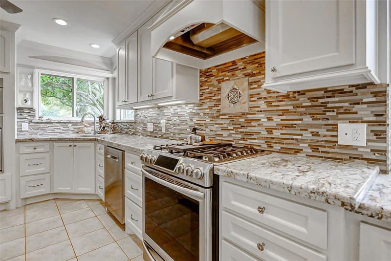 Kitchen corner with a stainless gas range under a white vent hood, granite counters, a mosaic tile backsplash, and a sink by the window