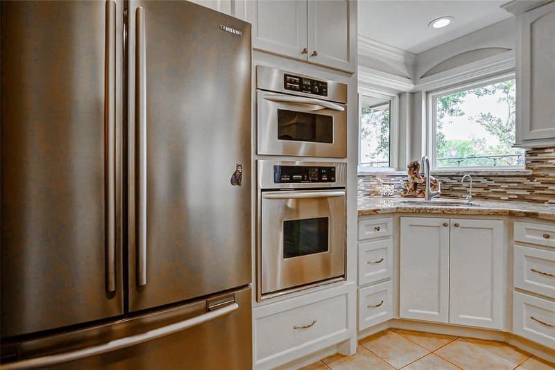 Stainless French-door refrigerator beside built-in stainless wall units, white cabinets, and a corner sink under two windows