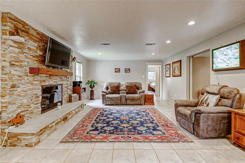 Living room with floor-to-ceiling stacked stone fireplace, wall-mounted TV, tile floor, recessed lights and a hall to a bedroom