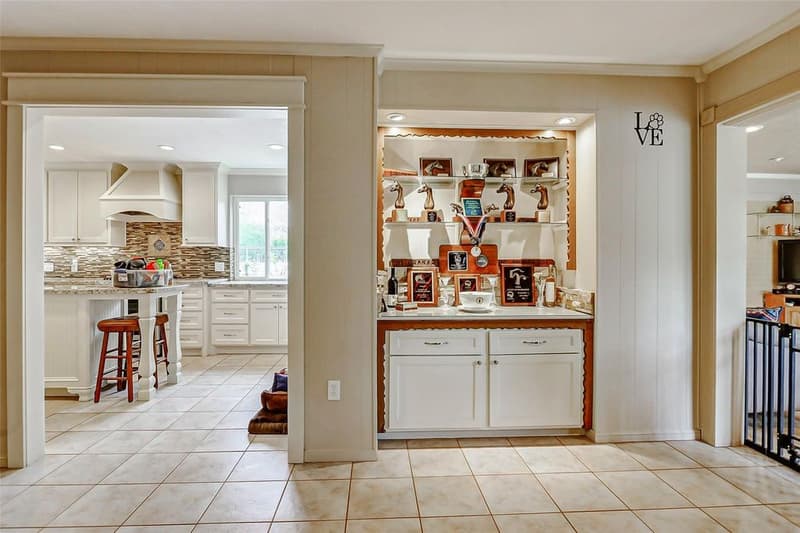 Built-in cabinet with lit shelves holding horse trophies and plaques, next to a doorway into a white kitchen with granite island