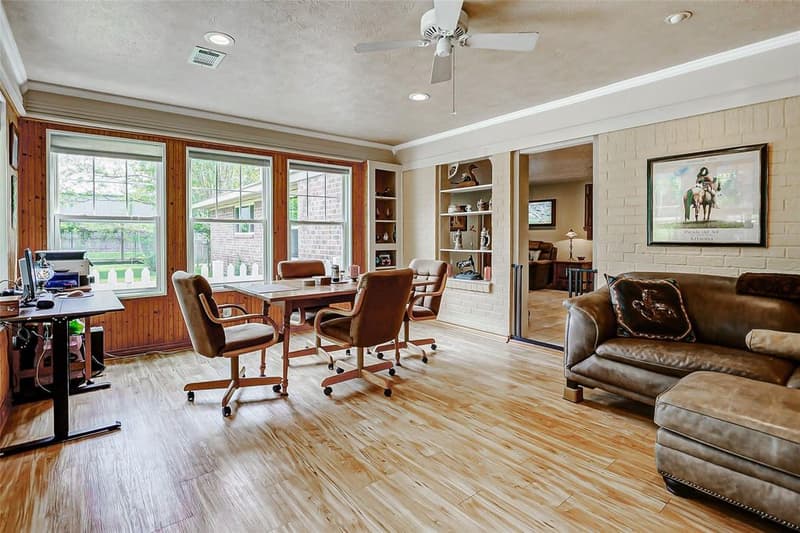 Wood-paneled room with three windows, wood-look floors, ceiling fan, built-in shelves, leather sofa and a desk area