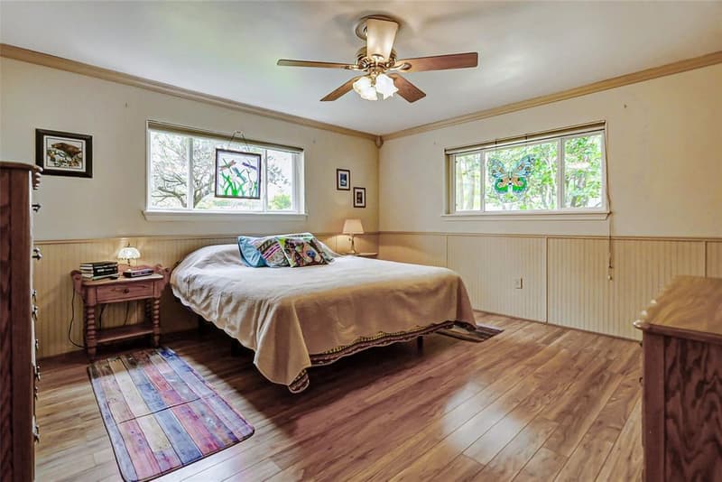 Bedroom with two windows, beadboard wainscoting, crown molding, wood-look floor and a ceiling fan with light