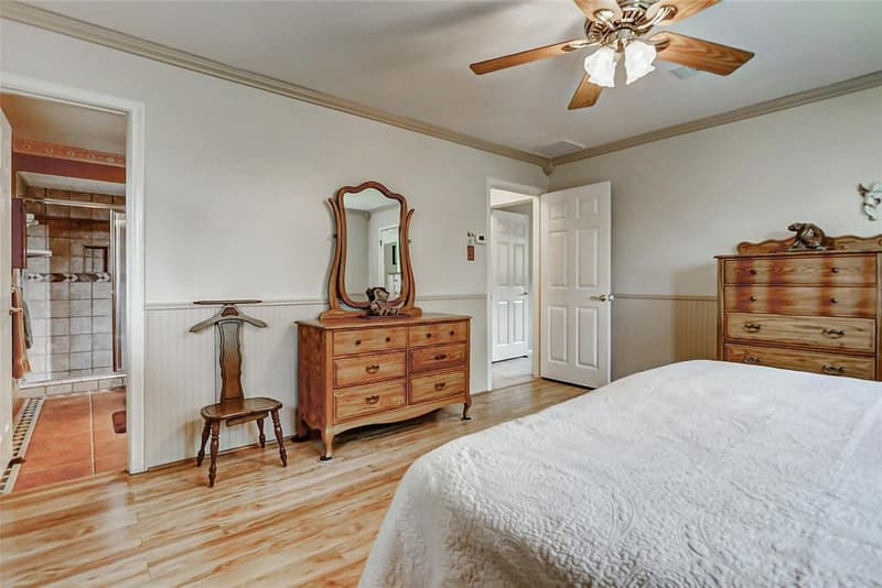 Bedroom with beadboard wainscoting, wood-look floor, ceiling fan and an open door to a bath with a tiled shower stall