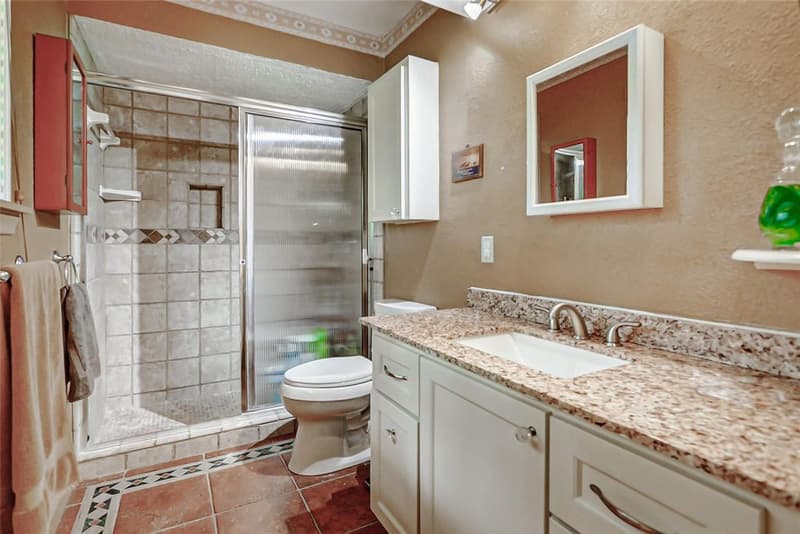 Bathroom with tiled shower stall, glass shower door, white vanity with granite countertop and terracotta-look tile floor