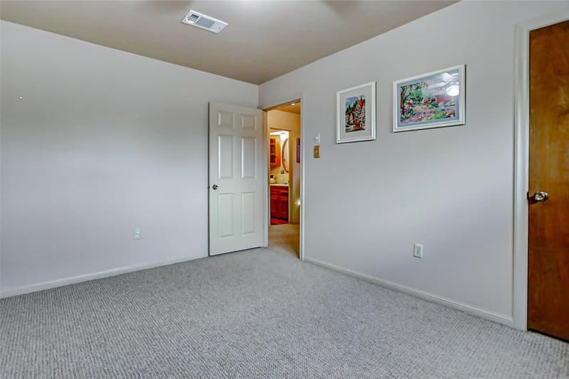Carpeted bedroom with light gray walls, framed art, and open door to a hallway with a bathroom beyond