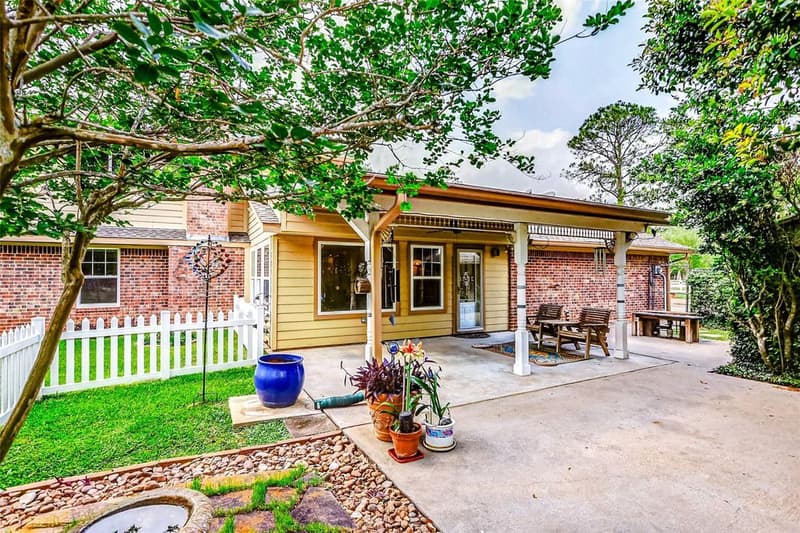 Covered back patio on a brick and siding home, with benches, potted plants, a white picket fence and shade trees