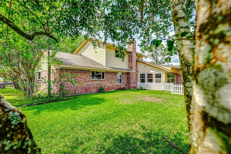 Two-story brick and siding home with chimney, wide green lawn, white picket fence and mature trees framing the view