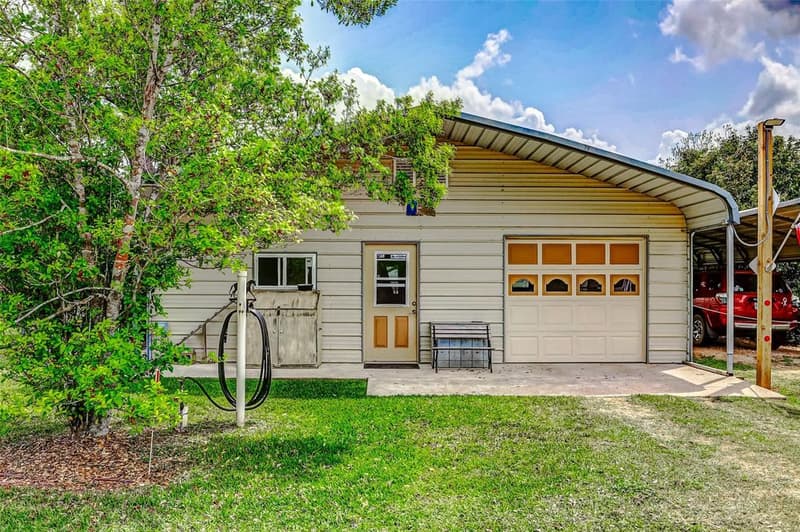 Cream metal outbuilding with a walk-in door, roll-up garage door and concrete apron, a metal carport beside it, under a leafy tree
