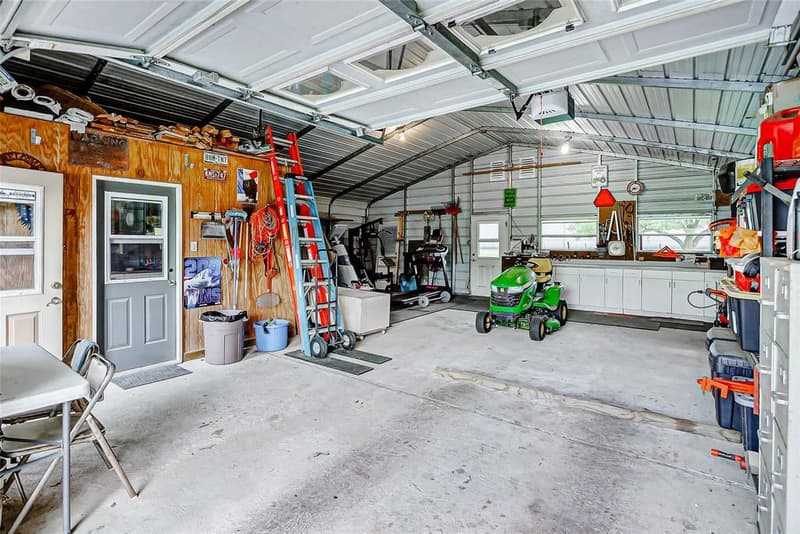 Inside a metal-framed workshop: concrete floor, riding mower, ladders, storage shelving, and a workbench with white cabinets