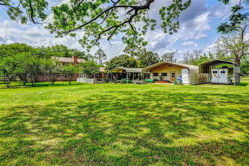 Wide lawn under overhanging tree branches looking toward a two-story brick home with chimney, metal carports, outbuilding and horse trailer